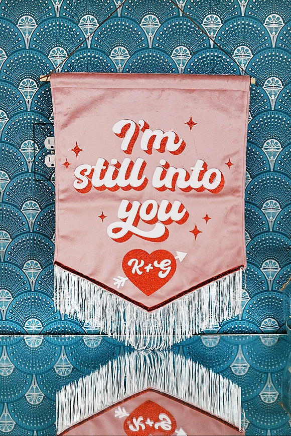 Wedding wall banner with glitter lettering and fringe trim hangs from a wooden dowel, set against patterned wallpaper with outlets visible