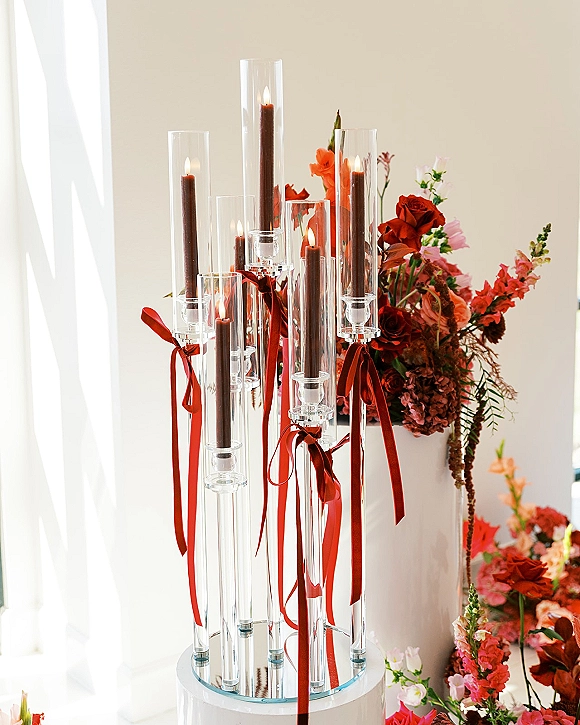 Wedding candle centerpiece with clear glass candle holders, red taper candles and ribbon bows beside roses in soft window light