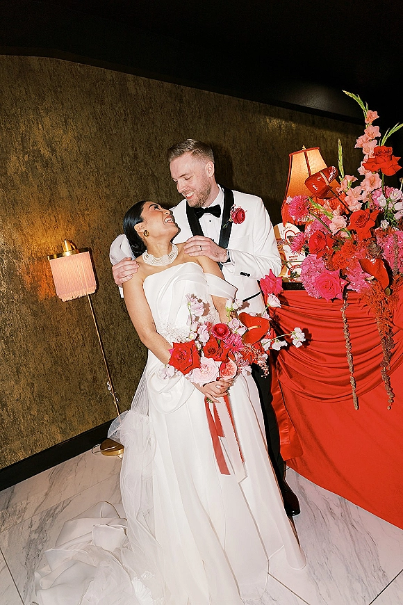 Couple portrait of bride and groom embrace, bride holding bouquet with red ribbons, against a textured wall on marble floor
