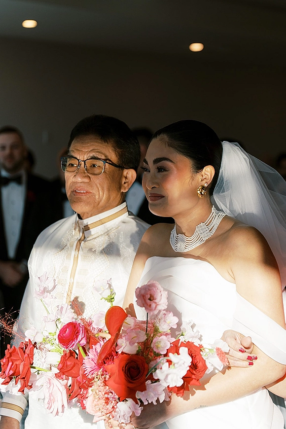Bride walking down aisle with father, holding a pink and red bouquet, veil and pearl necklace glowing under ceiling lights among guests