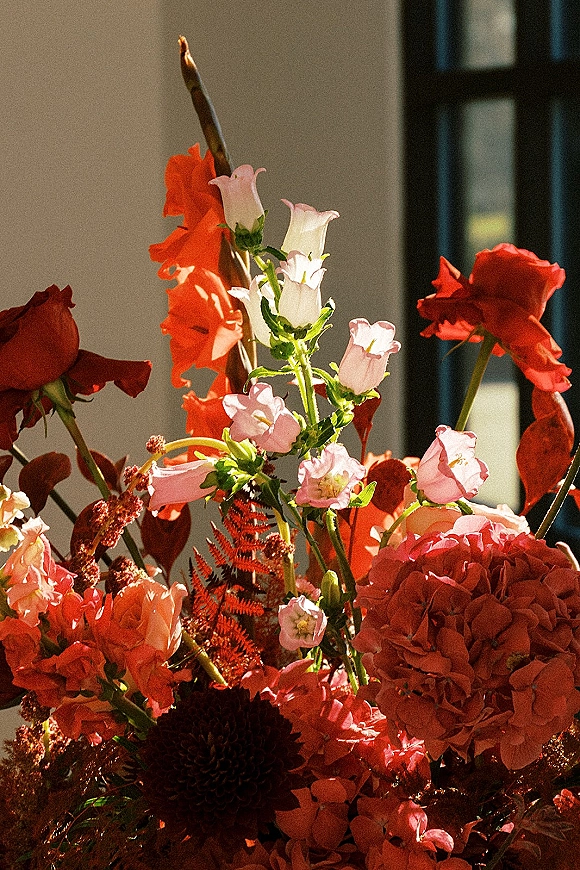 Wedding floral arrangement with red and pink wedding flowers, featuring roses, dahlias, carnations, and fern greenery in window light indoors