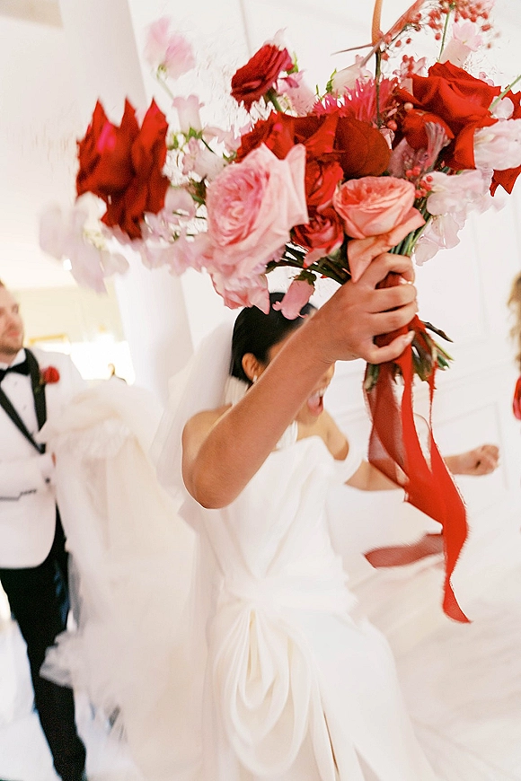 Bouquet toss moment as the bride throws a red and pink rose bouquet with long ribbons, veil flying, guests reaching in a white-walled venue