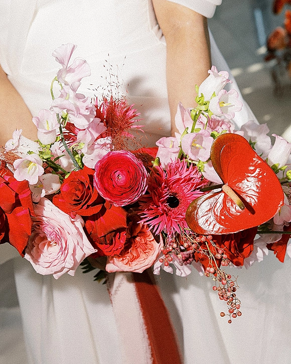 Bridal bouquet with red and pink bouquet blooms, featuring red anthurium, roses, and ranunculus, held over sunlit pavement shadows