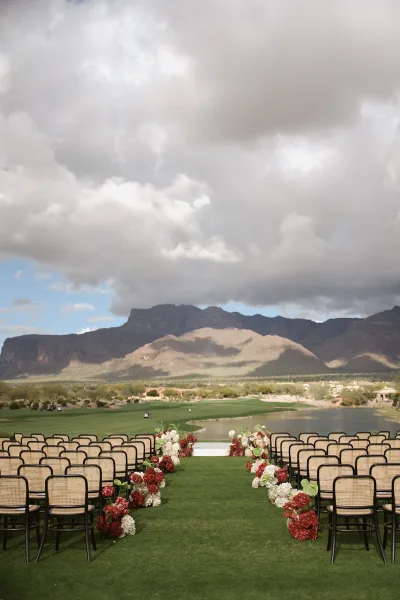 Ceremony aisle design with red and white aisle florals lining a grass walkway, cane-back and black chairs facing a white platform by mountains and lake