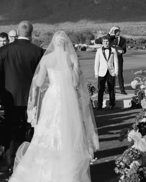 Ceremony moment as bride in lace dress and long veil approaches groom in white jacket and bow tie on a hilltop lawn stage