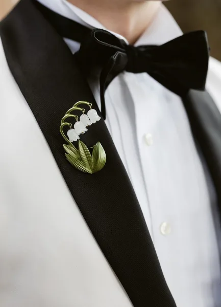 Groom boutonniere with lily of the valley flowers and green leaves pinned to a black satin lapel on a white tuxedo jacket indoors