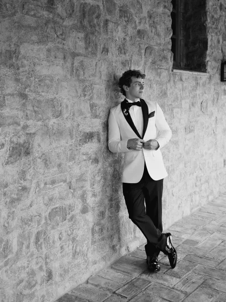 Groom portrait in a white tuxedo jacket with black bow tie, boutonniere and pocket square, leaning by a stone wall corridor walkway near a window