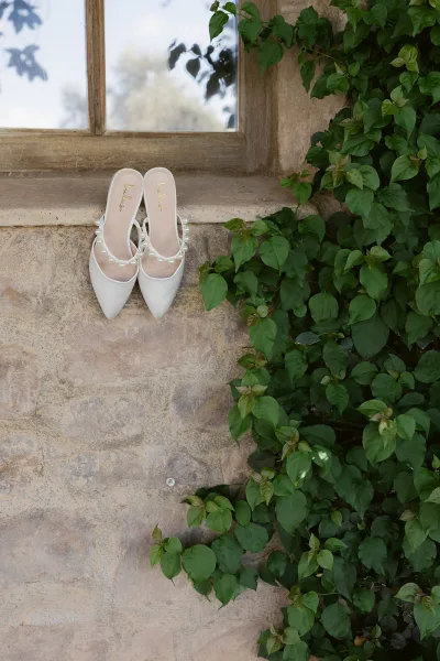 Wedding shoes with pearl strap embellishments, white pointed-toe heels resting on a stone window ledge framed by ivy vines