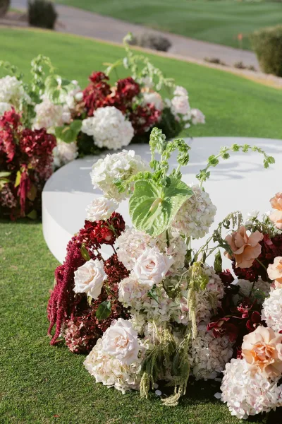 Wedding ceremony altar with a white circular stage, hydrangea and rose florals in burgundy and blush on a grass lawn path