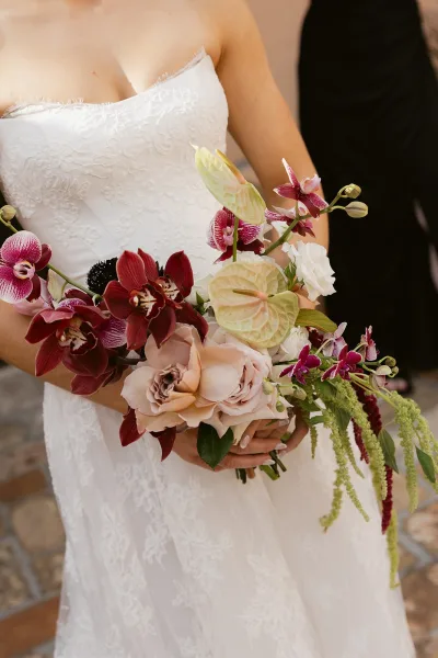 Bridal bouquet with tropical bridal bouquet orchids, anthurium, blush roses and trailing amaranthus against a strapless lace dress on stone pavement