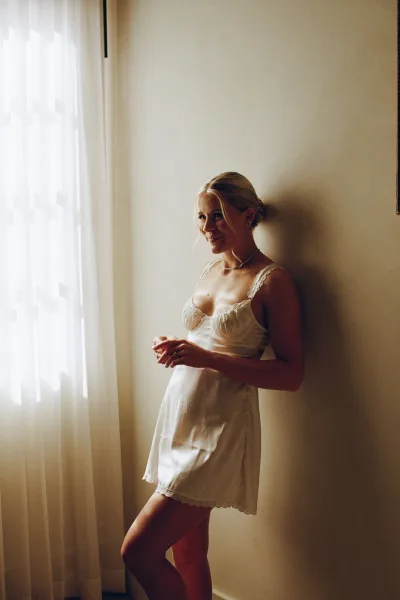Bridal portrait of a bride in a satin slip with lace trim, pearl necklace and wedding ring, posed by window light in a bedroom