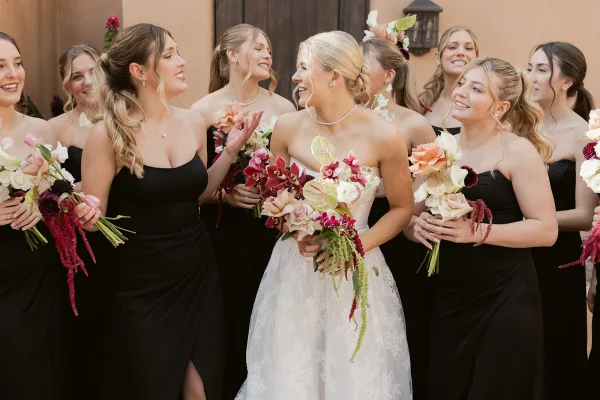 Bridesmaid group photo with bride with bridesmaids holding mismatched bouquets, posed by a stucco wall and wooden door with lantern
