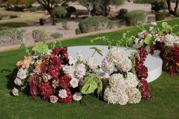 Wedding ceremony altar on a round white platform framed by hydrangeas, roses, anthurium, and burgundy florals on a garden lawn