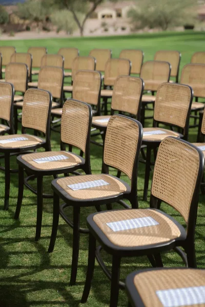 Ceremony seating with woven cane and black chairs lined in rows, reserved seat cards placed on each chair on a grassy lawn by trees