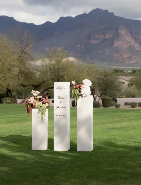 Wedding welcome sign acrylic on white pedestal plinths with hydrangea and hanging amaranthus on a lawn with mountains under cloudy sky