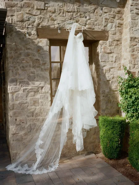 Wedding dress, lace wedding dress hanging from a wooden door with a long veil, framed by string lights against a stone wall