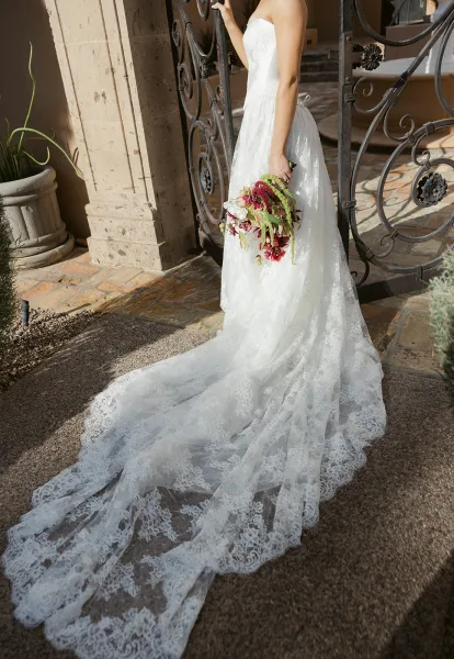 Bridal portrait of a bride in a strapless lace wedding dress with long train, holding a cascading red bouquet by a wrought iron gate under a stone archway