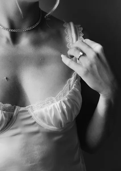 Engagement ring with emerald cut diamond on manicured hand, paired with lace lingerie and choker necklace against a dark backdrop