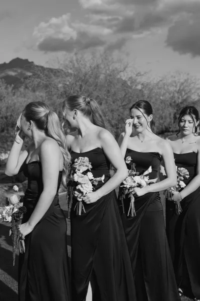 Bridesmaid portrait in black bridesmaid dresses holding bouquets, posed on grass with trees, mountains, and a cloudy sky backdrop