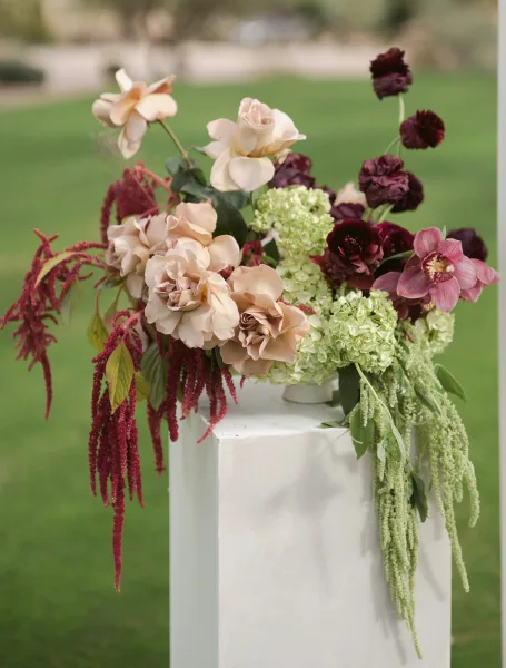 Wedding floral arrangement on a white pedestal plinth with roses, orchids, hydrangea and cascading amaranthus on a green garden lawn