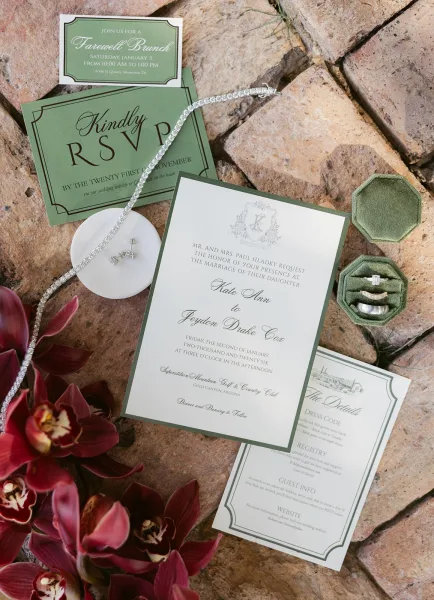Invitation suite with green wedding invitations, RSVP and details cards, rings and jewelry beside orchids on a stone surface flat lay