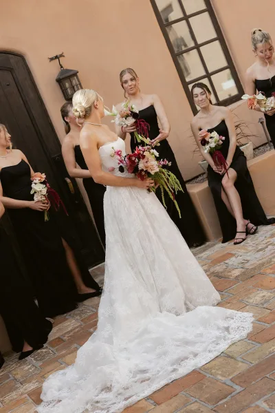 Bride with bridesmaids in black dresses holding burgundy and blush bouquets, posed by a stucco wall and wooden door in a courtyard.