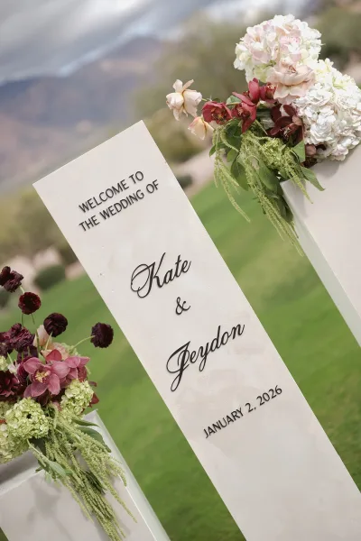 Wedding welcome sign with raised lettering on a wooden easel, framed by hydrangeas and orchids on an outdoor lawn with blurred hillside