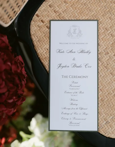 Wedding ceremony program with calligraphy typography and monogram crest beside a black high heel on a woven straw mat with greenery