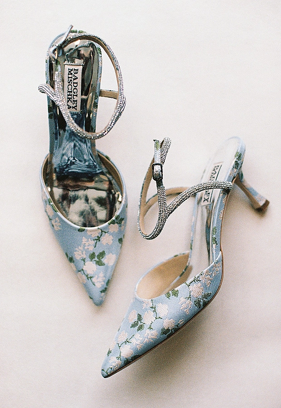 Bridal shoes in blue satin with floral fabric, pointed toes, ankle straps, and rhinestone buckle details on a white surface
