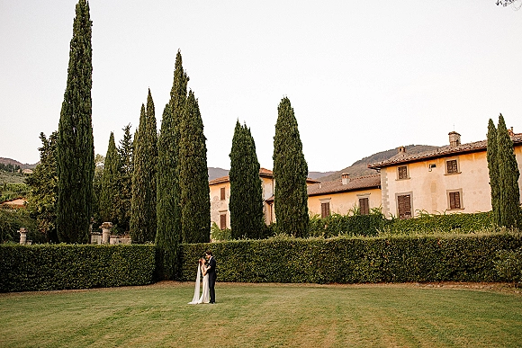 Couple portrait of bride and groom hugging, her long veil trailing on an estate lawn with cypress trees, villa and mountains behind