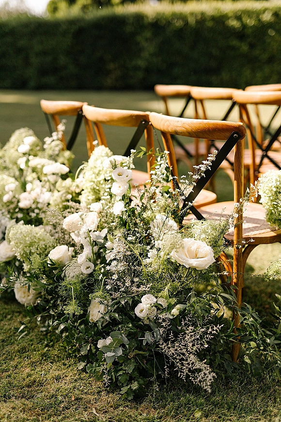 Ceremony aisle decor with grounded aisle flowers of white hydrangeas and roses, lush greenery, and wooden cross-back chairs on a lawn by hedges
