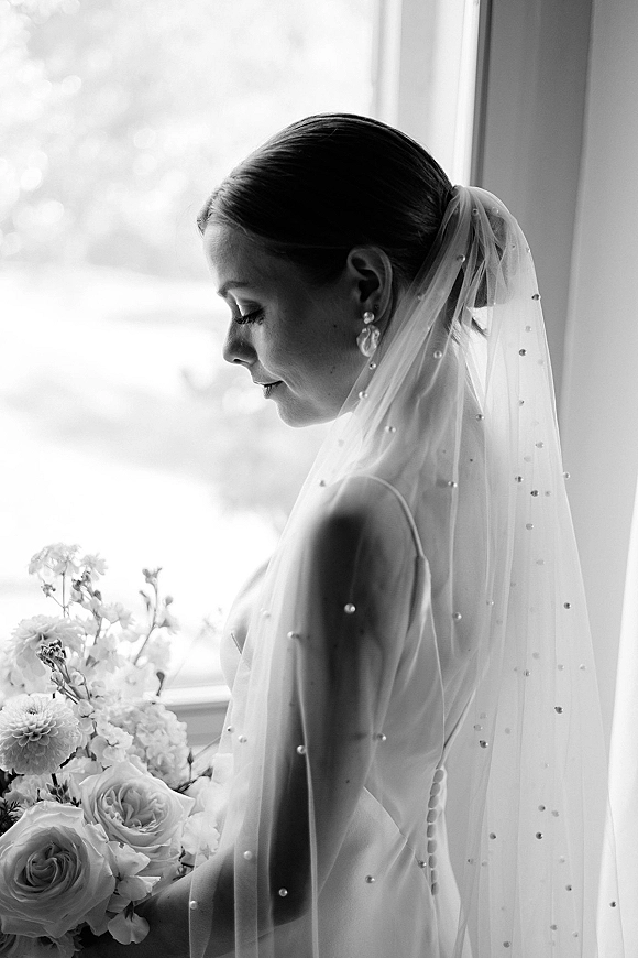 Bridal portrait in black and white of a bride in profile by a window, veil over her face, pearl earrings and rose bouquet in hand