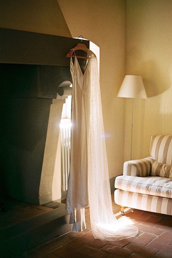 Wedding dress hanging on a wooden hanger with a lace veil, draped by a fireplace in a warm neutral room with terracotta tile floor