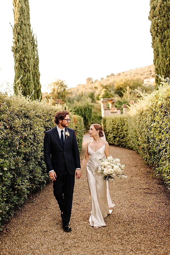 Couple portrait of bride and groom walking hand in hand, bride holding white bouquet on a gravel garden path with cypress trees behind