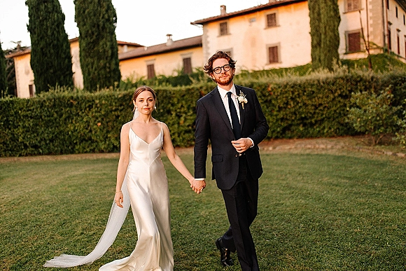 Couple portrait of bride and groom walking hand in hand, her chapel-length veil trailing, on a villa lawn with cypress trees