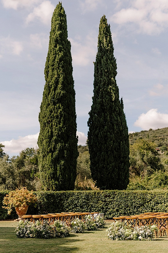 Ceremony setup for an outdoor wedding ceremony with wood chairs and green-white ground florals lining an aisle on a lawn beneath cypress trees