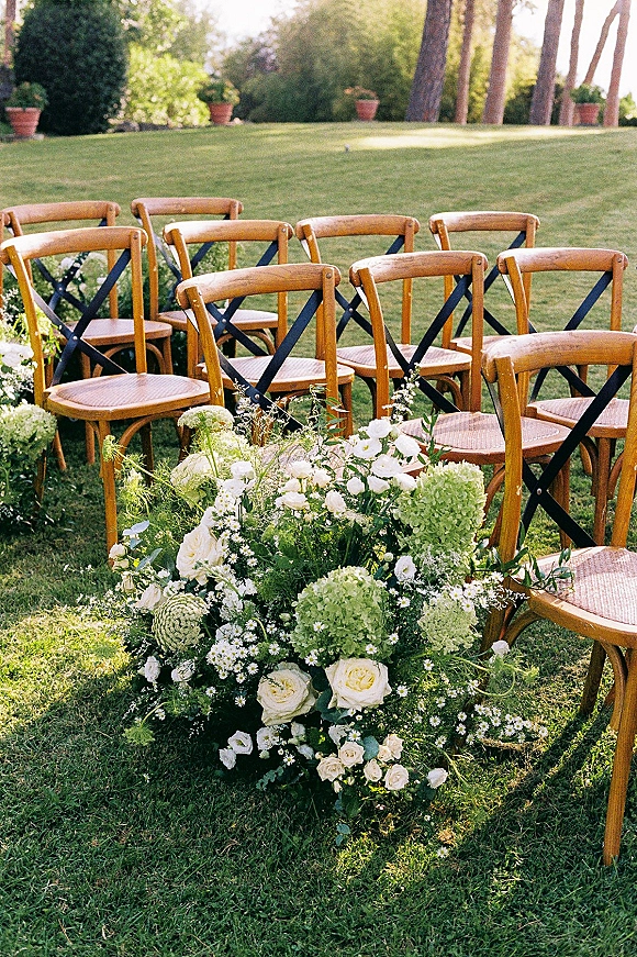 Ceremony seating with outdoor ceremony chairs lined along a floral aisle of white roses, hydrangeas, daisies, and greenery on a sunny lawn