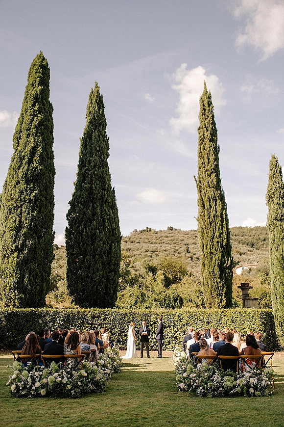 Outdoor wedding ceremony with bride and groom at the altar, long veil and white-green aisle flowers, framed by cypress trees and hedges