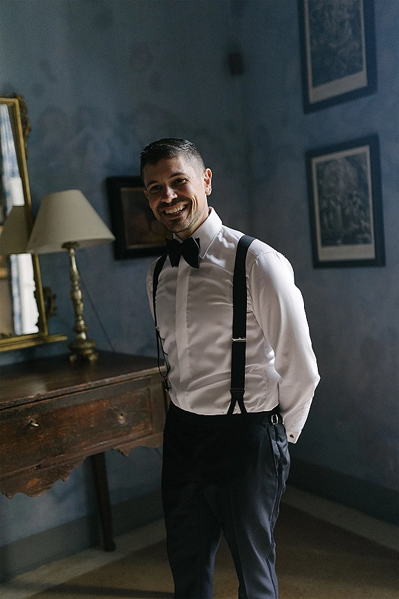 Groom portrait in suspenders, wearing a black bow tie and cufflinks, standing by a vintage dresser with mirror on a blue wall