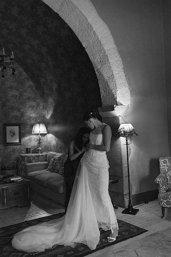 Bride getting ready in a lace wedding dress train, adjusting her necklace in a stone-arch living room with chandelier and lamps