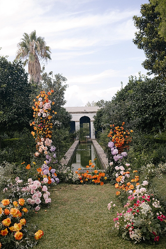 Ceremony aisle decor with a floral meadow of roses and wildflowers lining a garden path by a reflecting pool and arched building