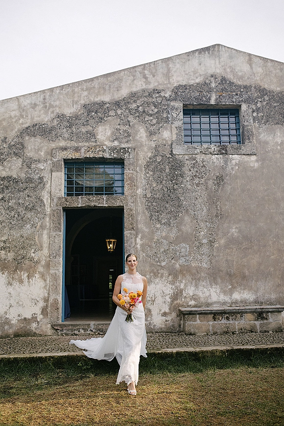 Bridal portrait of a bride holding bouquet with orange and pink flowers, in a strapless lace wedding dress by an arched stone doorway
