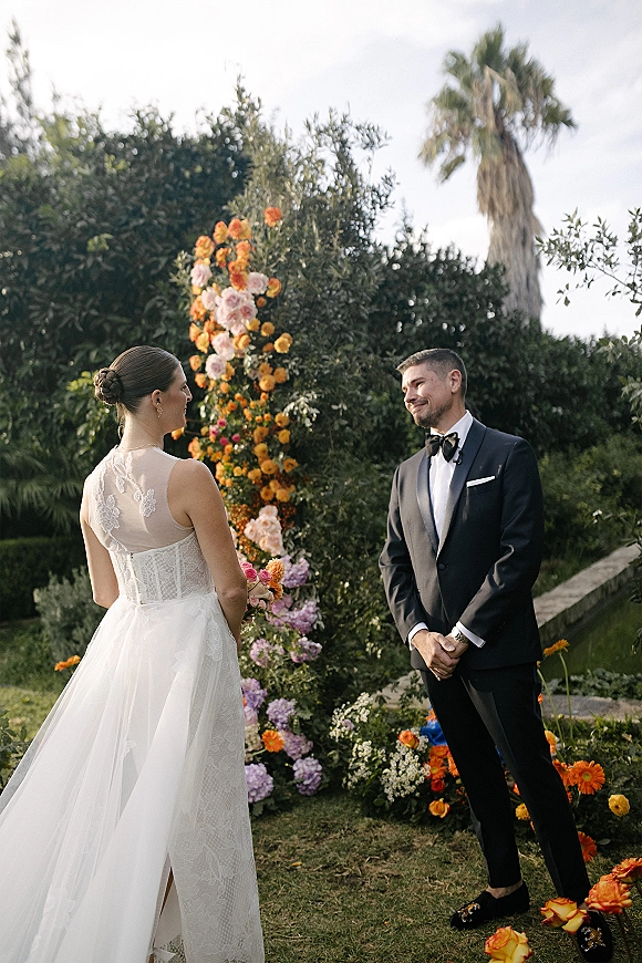 Wedding vows during outdoor wedding ceremony as bride in lace bodice with bouquet faces groom in tux under floral arch in lush garden greenery