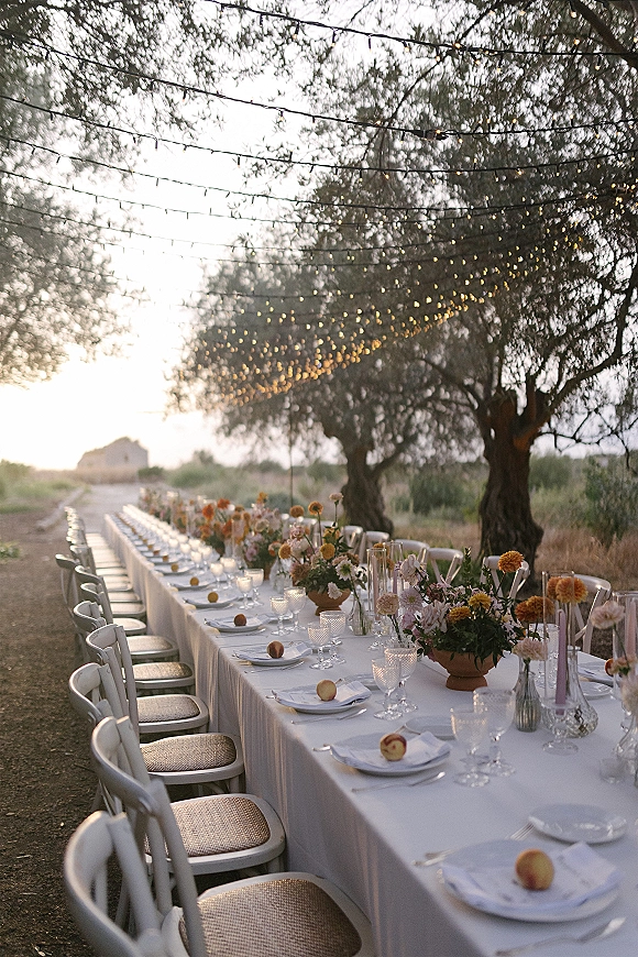 Reception tablescape with long banquet table wedding setup, white linens, floral centerpieces, taper and votive candles under string lights at sunset outdoors