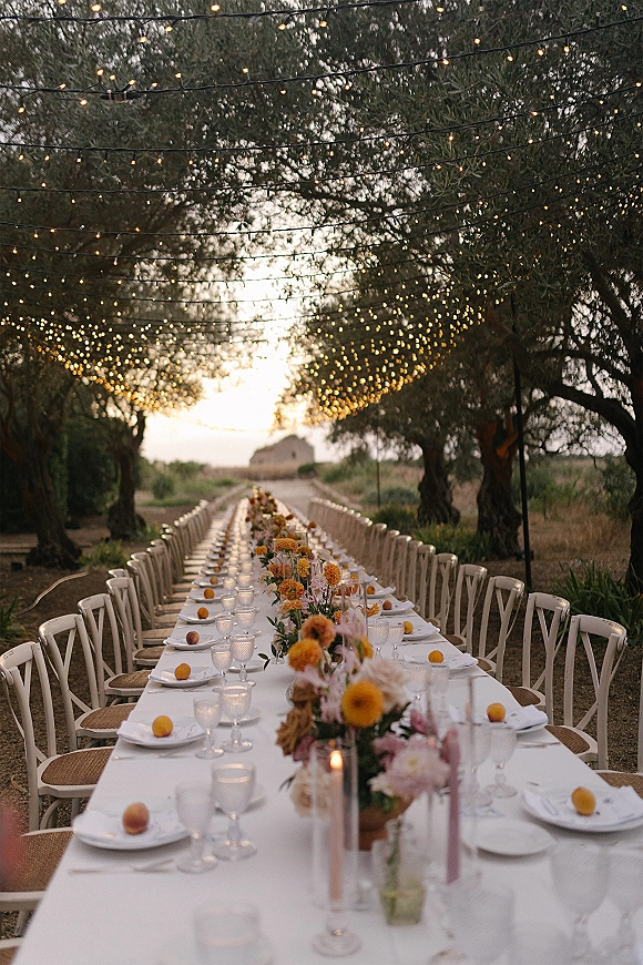 Reception tablescape with an outdoor banquet table in white linen, floral centerpieces, taper candles, and string lights under trees
