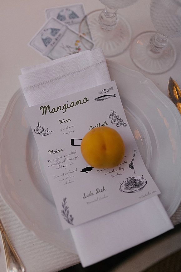 Wedding place setting with a wedding menu card atop a white plate, linen napkin, orange accent, silverware, and champagne coupes on white cloth