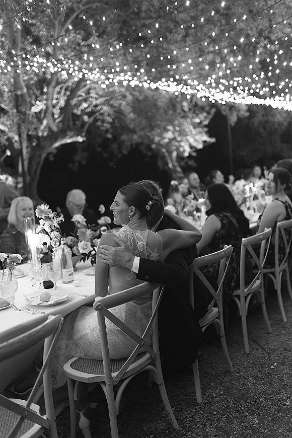Reception moment as newlyweds seated together beneath string lights, bride in lace dress hugging groom at candlelit garden dinner tables