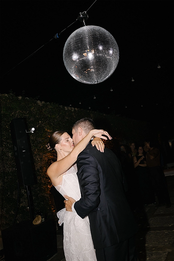 First dance as bride in a strapless lace wedding dress and groom in tuxedo slow dance under string lights on a stone patio at night