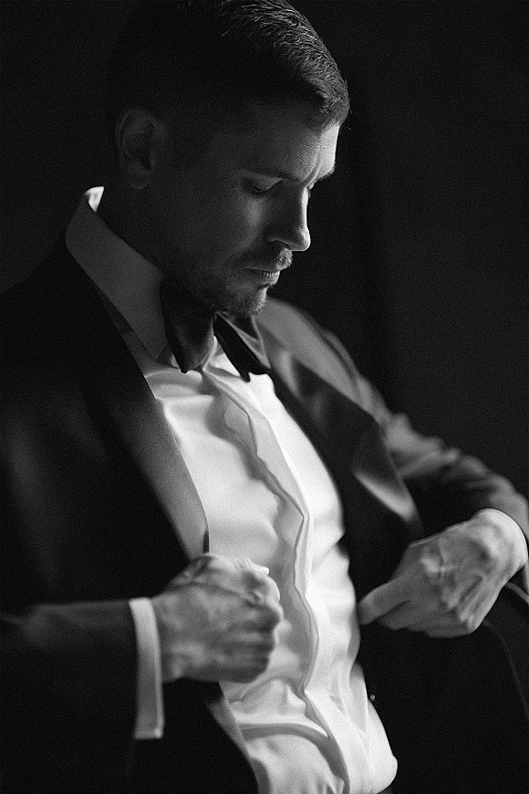 Groom portrait in moody shadows, black tie groom adjusting his tuxedo jacket with bow tie and cufflinks in a dark interior