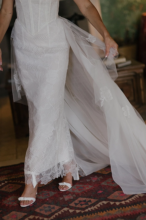 Wedding dress details show a lace wedding dress with corset bodice, tulle overskirt, and bow bridal sandals on a vintage rug indoors